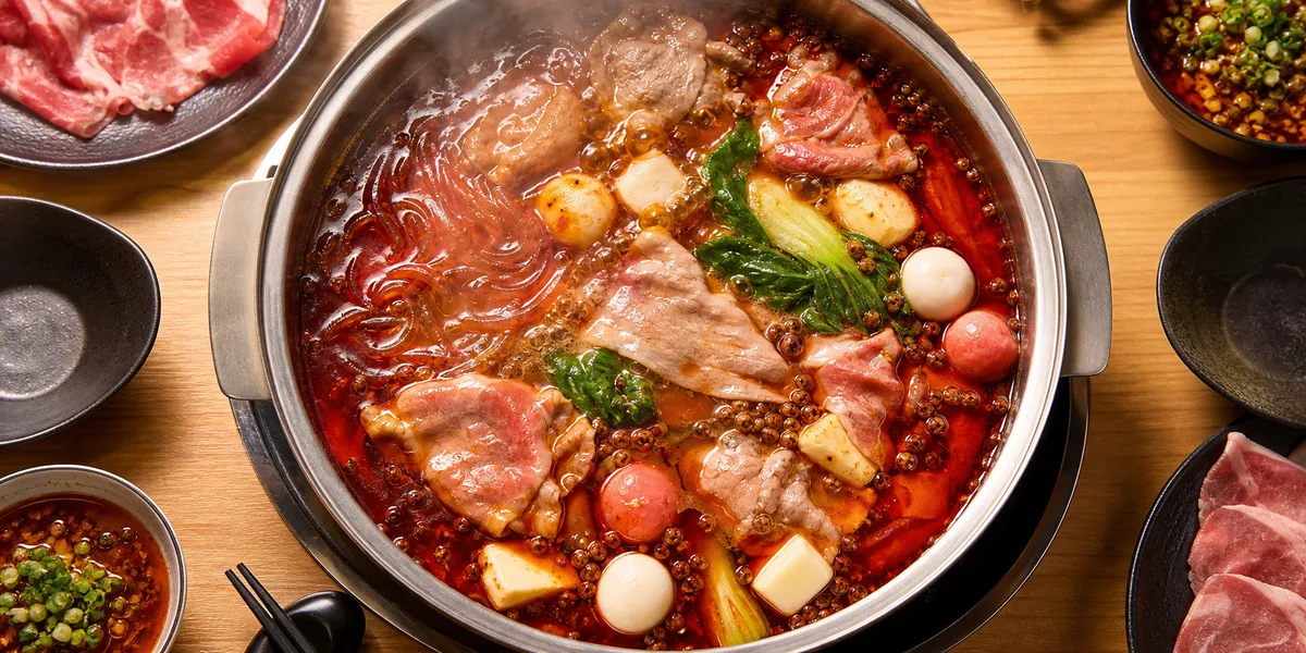 Overhead view of a Shabu-yo-style hot pot with red malatang broth bubbling, lamb, kohaku dango, and glass noodles floating on the surface